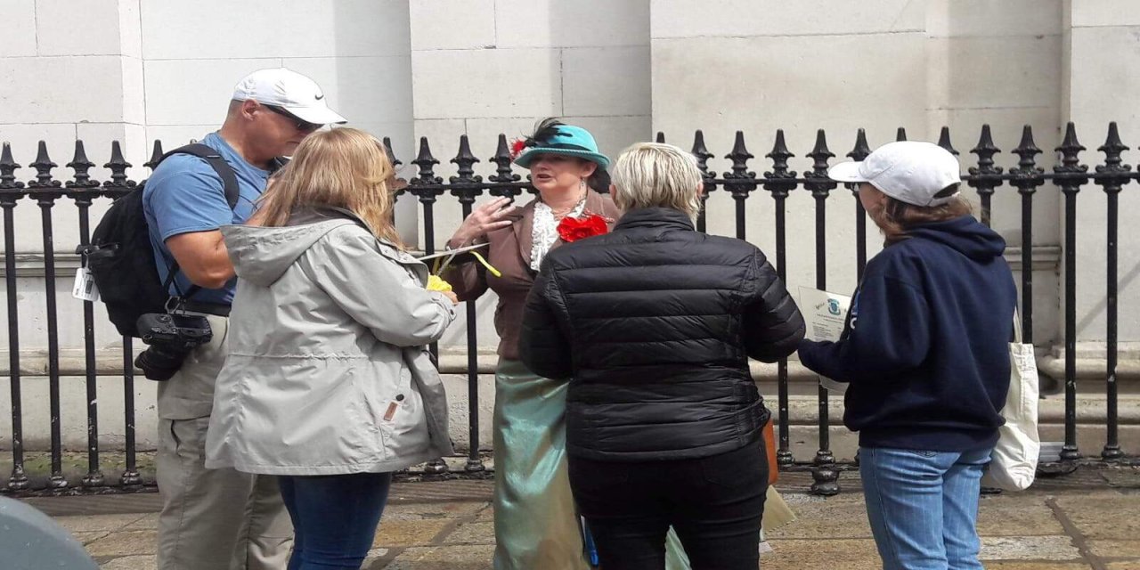 4 tourists standing and listening to tour guide singing. Standing infront of black gates and white building. 